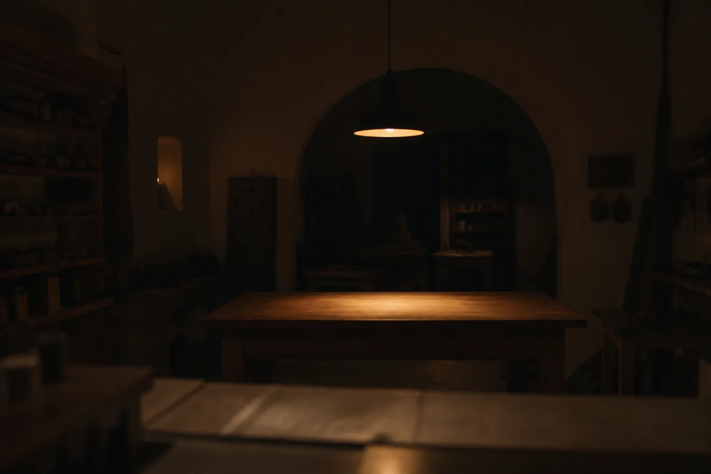 An empty workshop interior at night, a single low pendant lighting a bare bench top while the room behind falls into deep shadow.